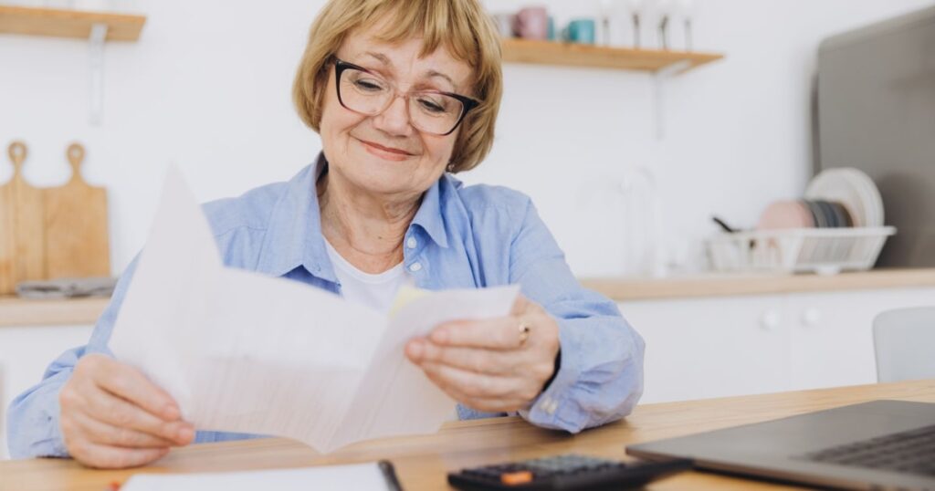 Une dame assise à une table de cuisine sourit en regardant un document papier. 